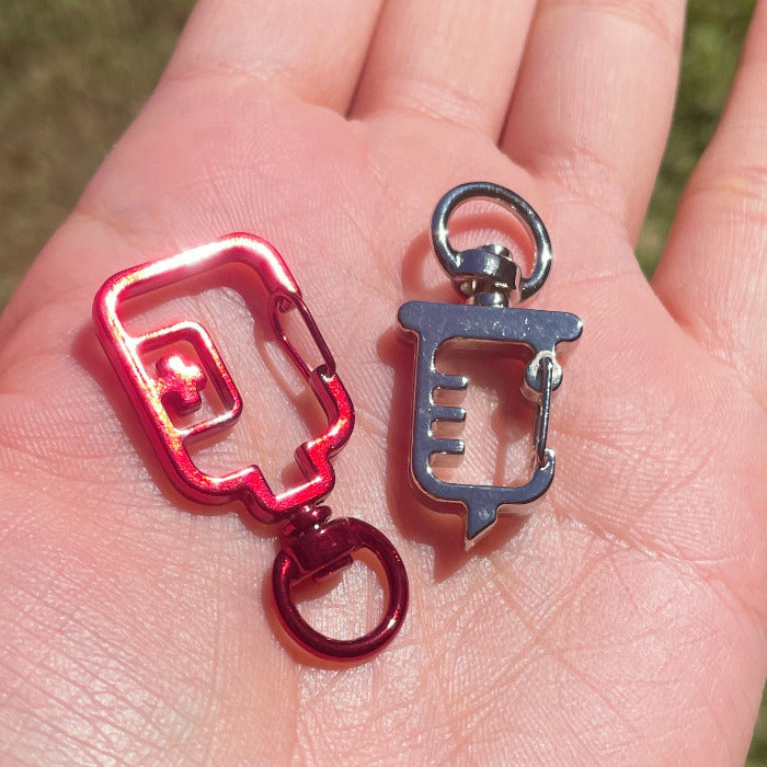 A red blood bag shaped keyring clasp and a silver syringe shaped keyring clasp laid in the palm of subject's hand