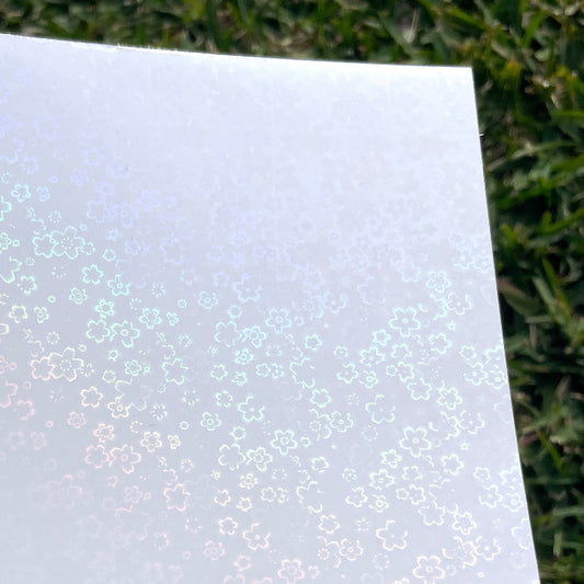 A sheet of the A4 flower holo glossy cold laminate lays on a patch of grass in direct sunlight. The design features various five petal flowers scattered across the page.