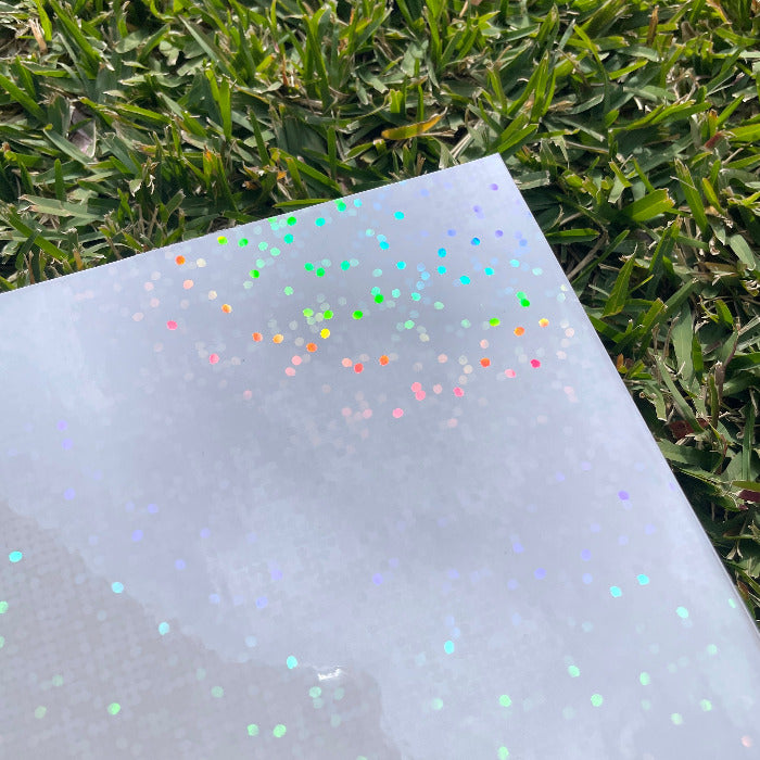 A sheet of the disco dots holo glossy cold laminate lays on a patch of grass in direct sunlight. The design features large holographic rainbow dots that dance in the light.