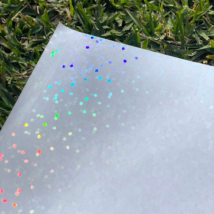 A sheet of the disco dots holo glossy cold laminate lays on a patch of grass in direct sunlight. The camera is looking down on the top left corner. The design features large holographic rainbow dots that dance in the light.