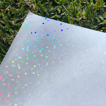 A sheet of the disco dots holo glossy cold laminate lays on a patch of grass in direct sunlight. The camera is looking down on the top left corner. The design features large holographic rainbow dots that dance in the light.