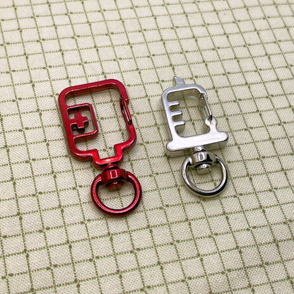 A red blood bag shaped keyring clasp and a silver syringe shaped keyring clasp laid flat on a grid background