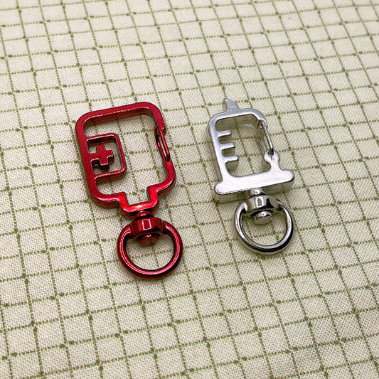 A red blood bag shaped keyring clasp and a silver syringe shaped keyring clasp laid flat on a grid background