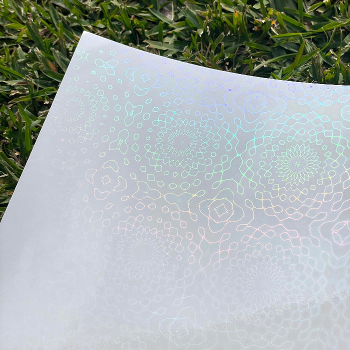 A sheet of the A4 bubble web holo glossy cold laminate lays on a patch of grass in direct sunlight. The Design features bubbles and loops that form a mandala-like spiral.