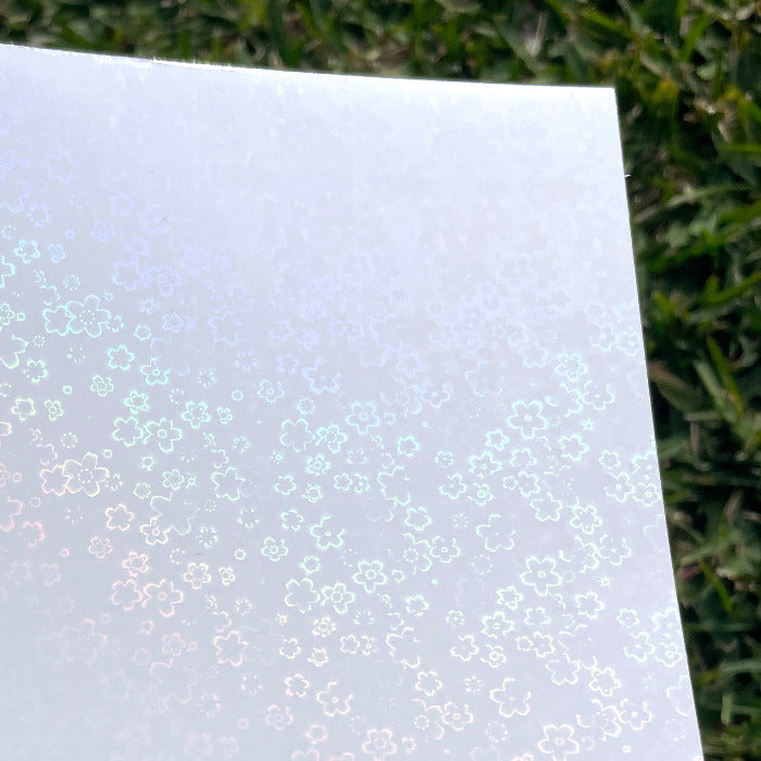 
A sheet of the A4 flower holo glossy cold laminate lays on a patch of grass in direct sunlight. The design features various five petal flowers scattered across the page.